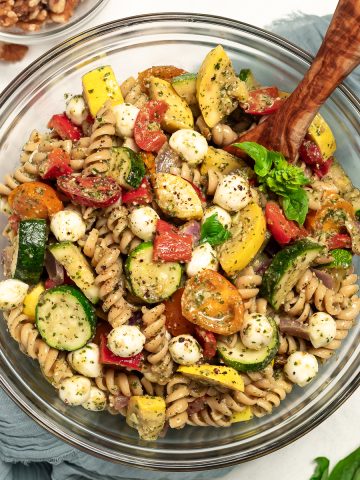 A bowl of walnut pesto pasta salad with roasted vegetables sits on a table with a napkin and a serving spoon. A bowl of walnuts sits off to the side.