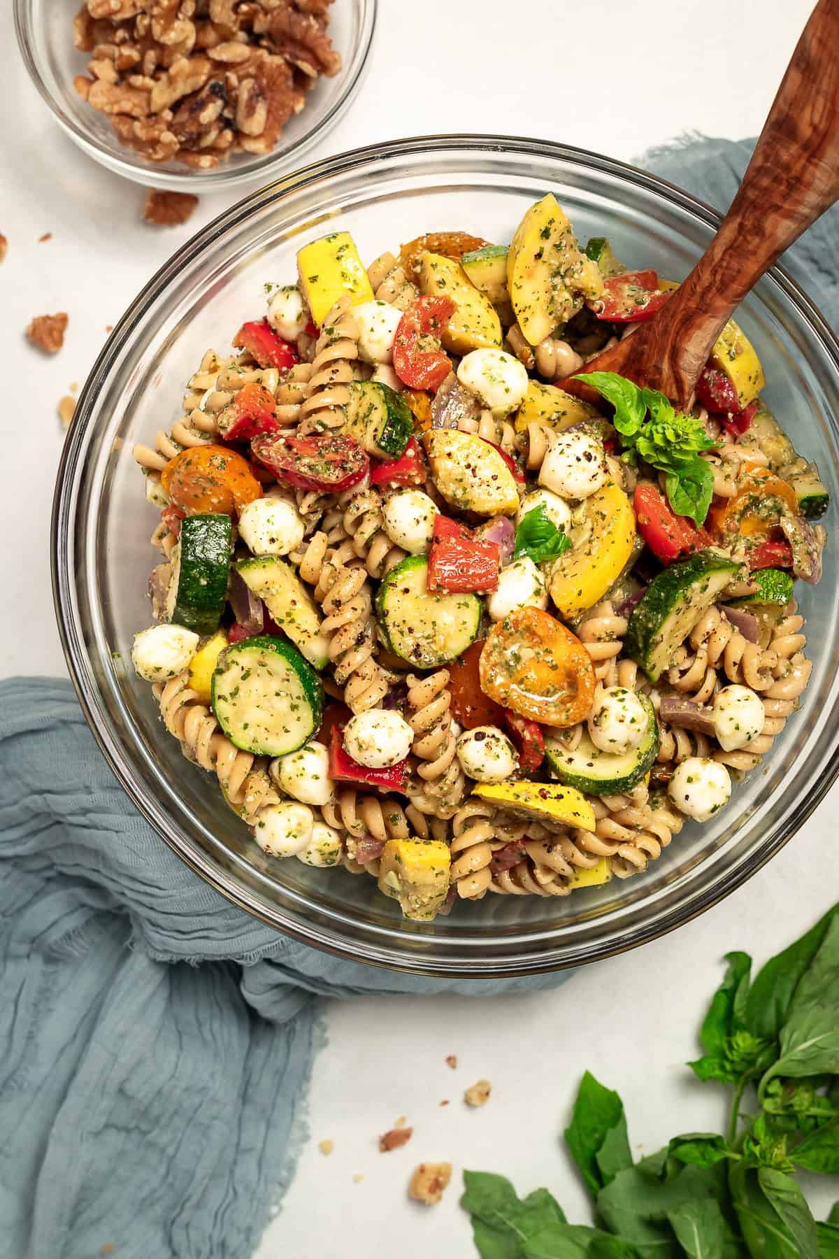 A bowl of walnut pesto pasta salad with roasted vegetables sits on a table with a napkin and a serving spoon. A bowl of walnuts sits off to the side.