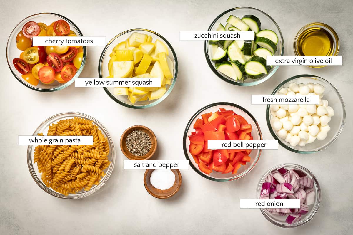 Ingredients for walnut pesto pasta salad with roasted vegetables. Cherry tomatoes, yellow summer squash, whole grain pasta, salt and pepper, zucchini, extra virgin olive oil, fresh mozzarella, red bell pepper, and red onion.