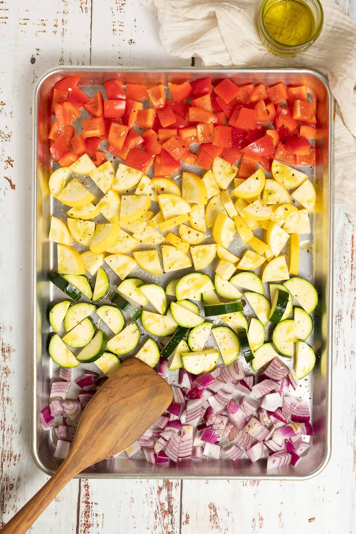 A pan of vegetables is ready to go in the oven. A spatula and a bowl of extra virgin olive oil sit nearby.