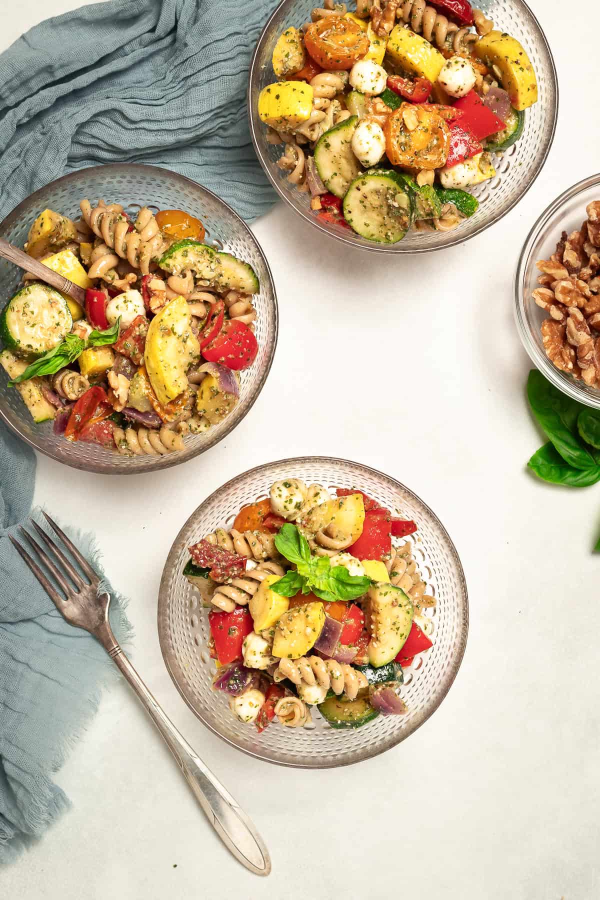 Three small bowls of pasta salad with roasted vegetables dressed with walnut pesto. Two forks lay on the table, and a bowl of walnuts sits off to the side.