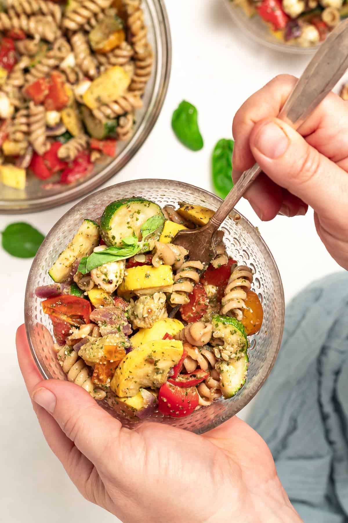 One hand holds a bowl of pasta salad with roasted vegetables and pesto. Another hand holds a fork that is digging in to the salad. A large bowl of salad sits on the table in the background.