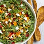 Arugula, sweet potato, farro, and pomegranate tossed together in a bowl with salad tossers sitting next to it.