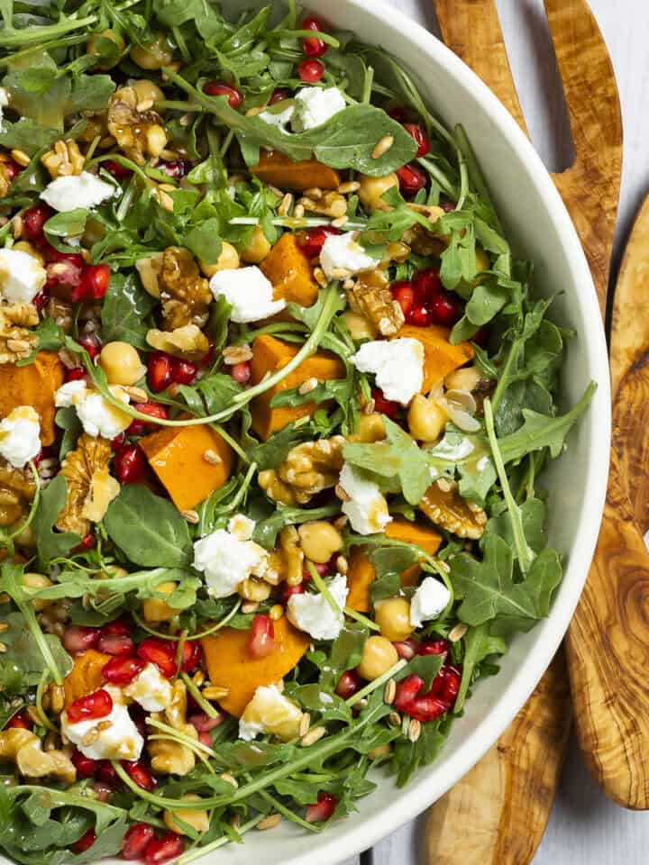 Arugula, sweet potato, farro, and pomegranate tossed together in a bowl with salad tossers sitting next to it.