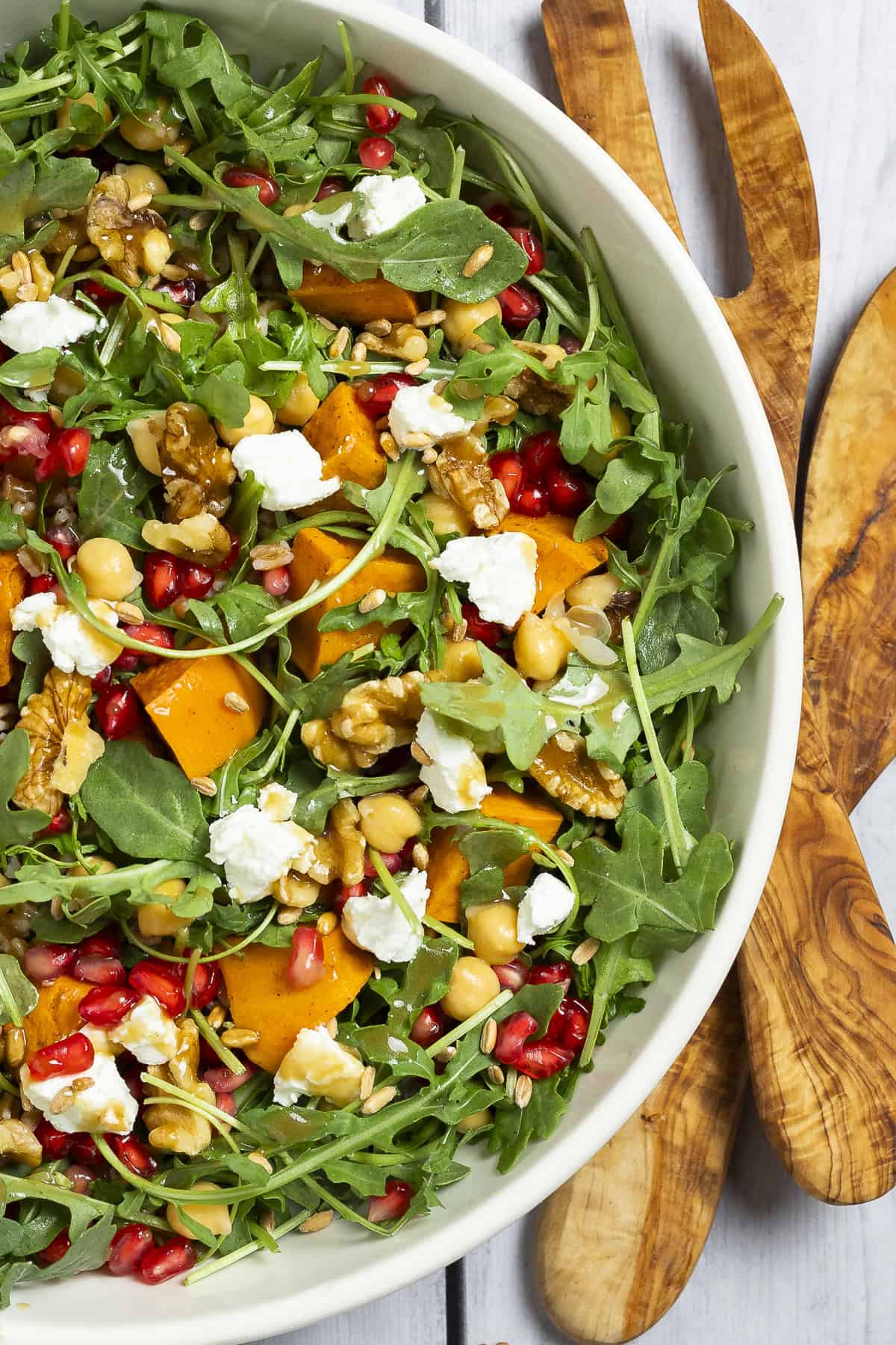 Farro and arugula salad in a white bowl with salad tossers. Garnished with sweet potatoes, pomegranate arils, walnuts, and goat cheese.