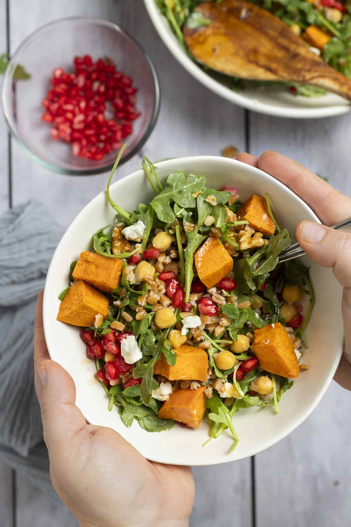 Someone holds a bowl of farro salad with arugula, sweet potatoes, pomegranate, chickpeas, and goat cheese. 