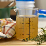 Turkey broth is shown in a measuring cup with a kitchen towel and herbs.