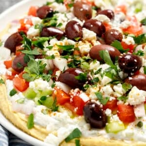 A plate filled with Greek layer dip including hummus, tzatziki, cucumbers, tomatoes, olives, shallot, and parsley.