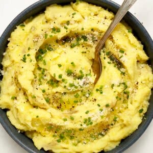 A bowl of fluffy mashed potatoes with Parmesan cheese and olive oil with a spoon. Garnished with chives, grated Parmesan, black pepper, and drizzled with olive oil.