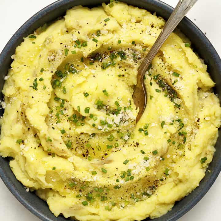 A bowl of fluffy mashed potatoes with Parmesan cheese and olive oil with a spoon. Garnished with chives, grated Parmesan, black pepper, and drizzled with olive oil.