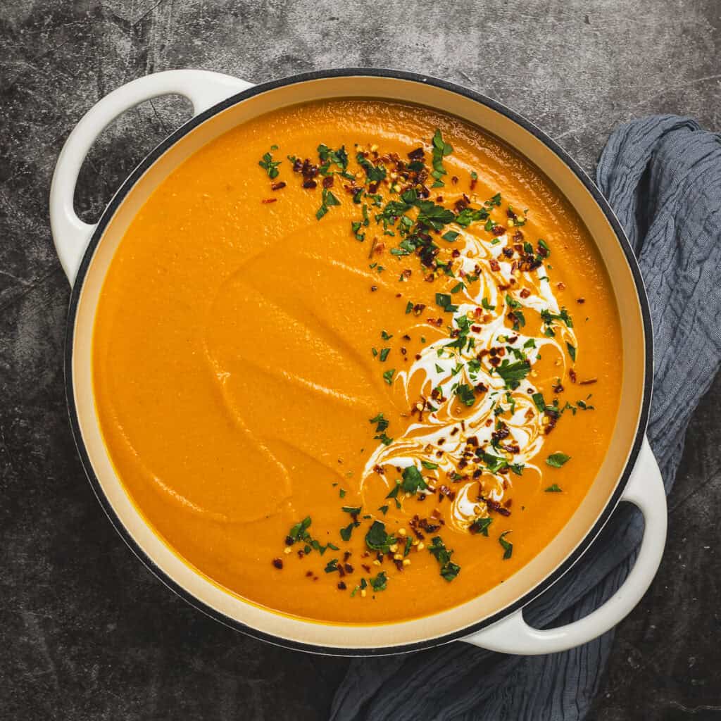 A bowl of carrot and red lentil soup in a pan, garnished with yogurt, fresh herbs, and red pepper flakes.