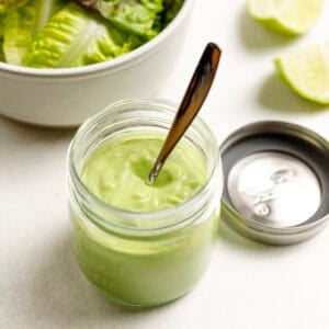 Cilantro avocado dressing in a jar with a bowl of lettuce and limes in the background.
