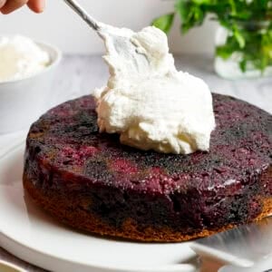 Blueberry Olive Oil Cake on a plate accompanied by a bowl of whipped cream. Someone is adding a dollop of whipped cream to the top of the cake.
