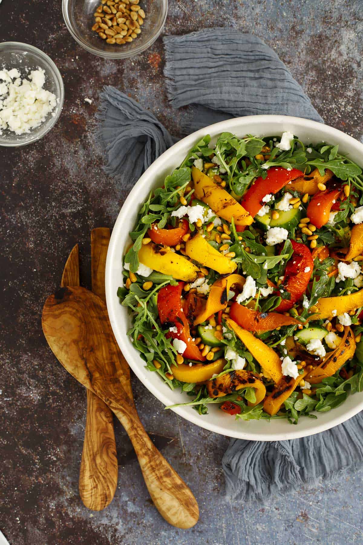 Grilled pepper salad in a bowl with salad servers. A bowl of feta cheese and a bowl of toasted pine  nuts sit nearby.