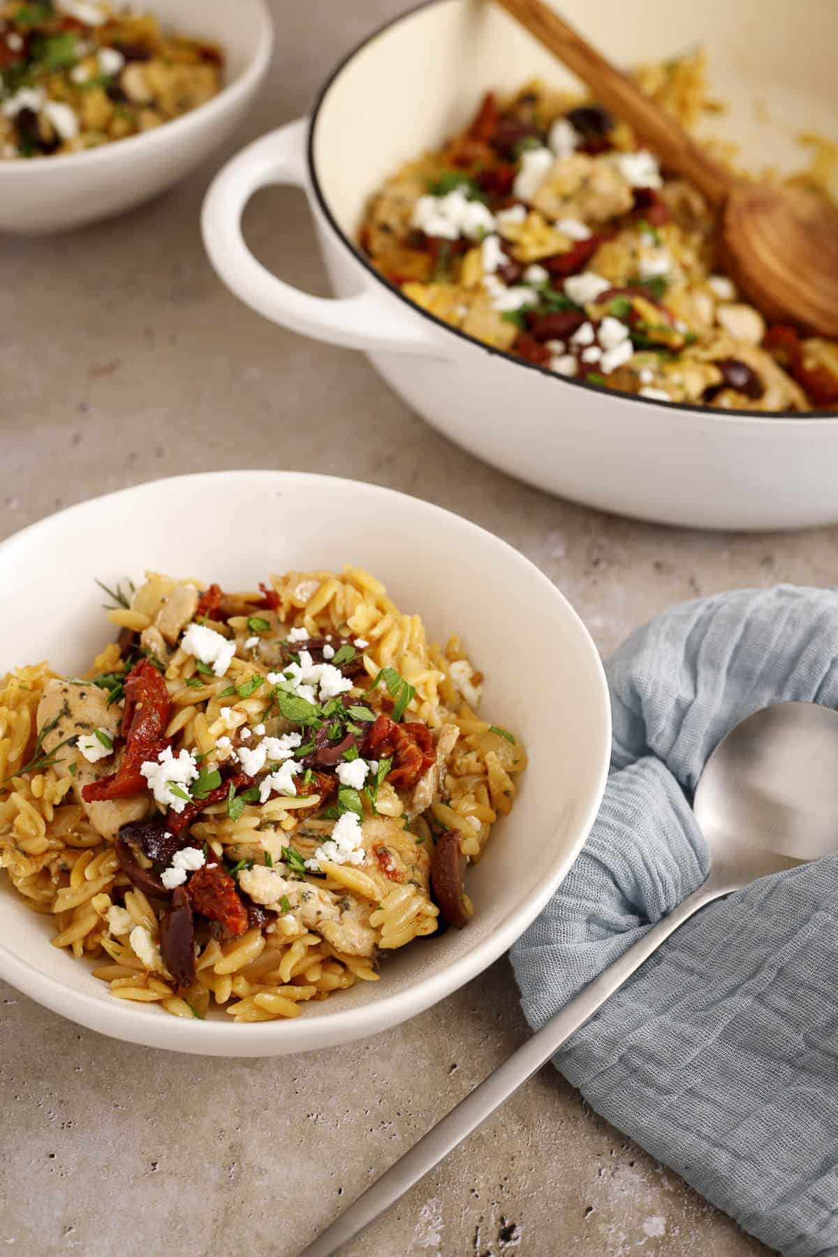 Mediterranean chicken orzo skillet in a pan with a spoon, with two bowls of it served nearby.