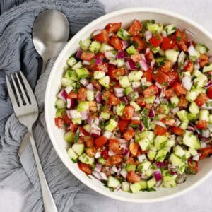 Mediterranean chopped salad in a bowl, featuring tomatoes, cucumbers, red onion, and herbs. The bowl is accompanied by a napkin and salad servers.