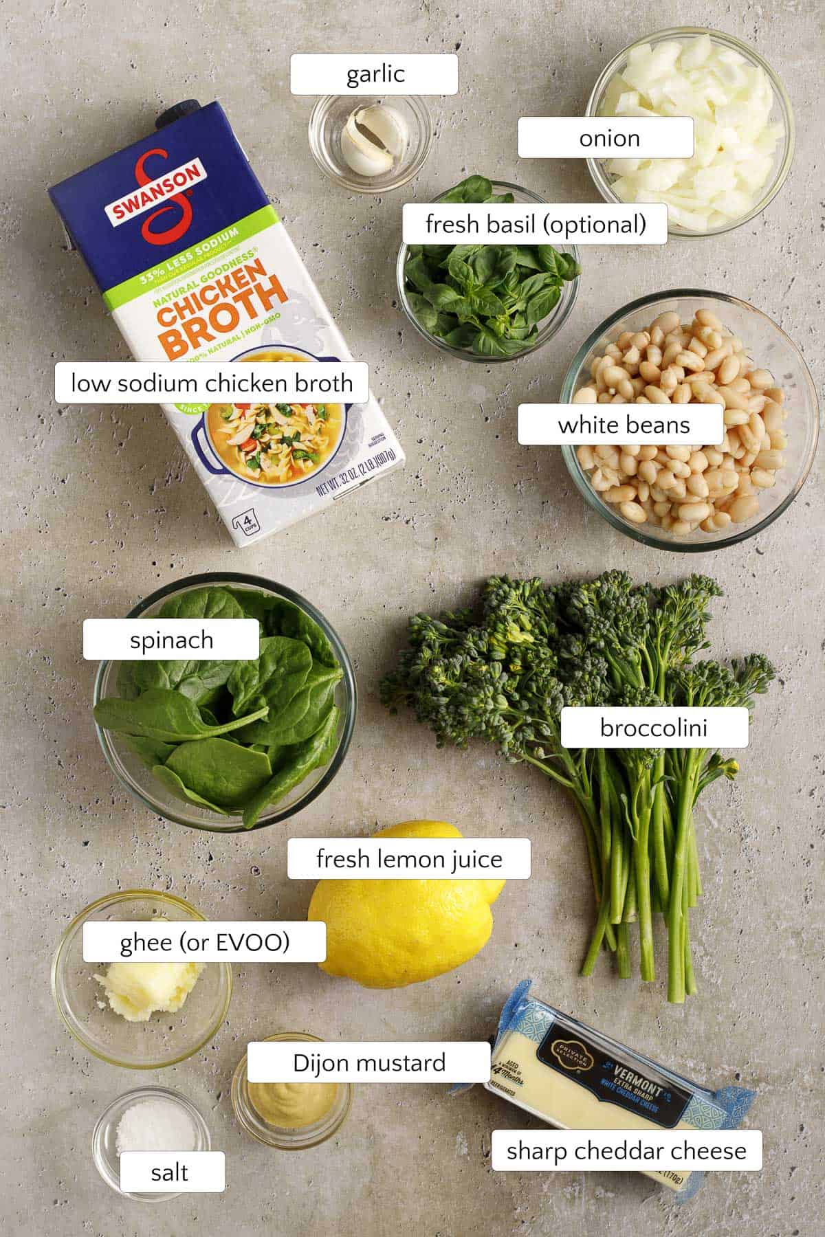 Labeled overhead image of ingredients for broccolini soup on a light countertop, including low sodium chicken broth, garlic, onion, fresh basil, white beans, broccolini, spinach, fresh lemon juice, ghee or EVOO, Dijon mustard, salt, and sharp cheddar cheese.