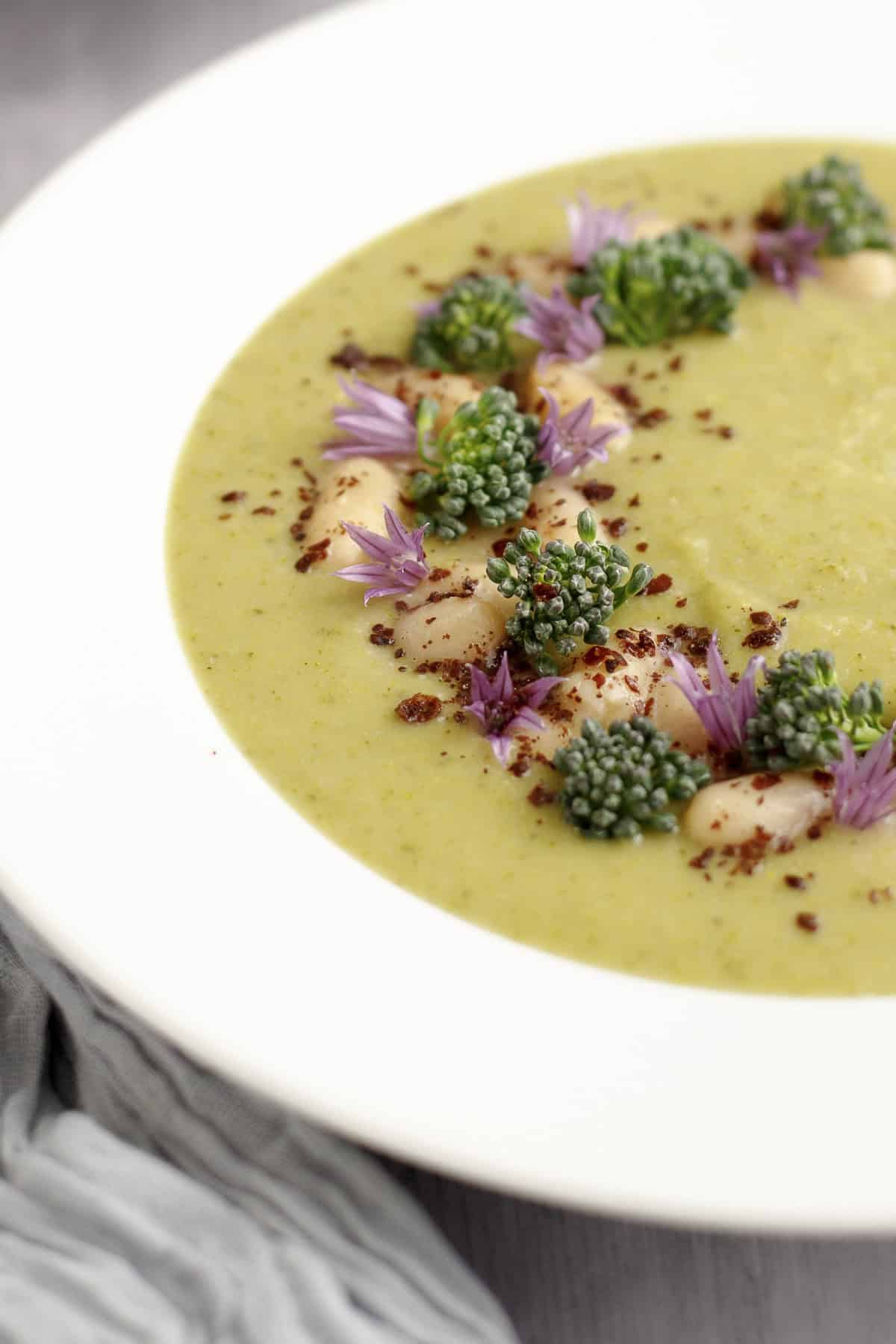 A close-up of a bowl of creamy broccolini soup garnished with white beans, broccolini florets, purple chive blossoms, and a sprinkle of sumac, served in a white bowl on a gray linen napkin.