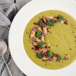 A close-up of a bowl of creamy broccolini soup garnished with white beans, broccolini florets, purple chive blossoms, and a sprinkle of sumac, served in a white bowl on a gray linen napkin.