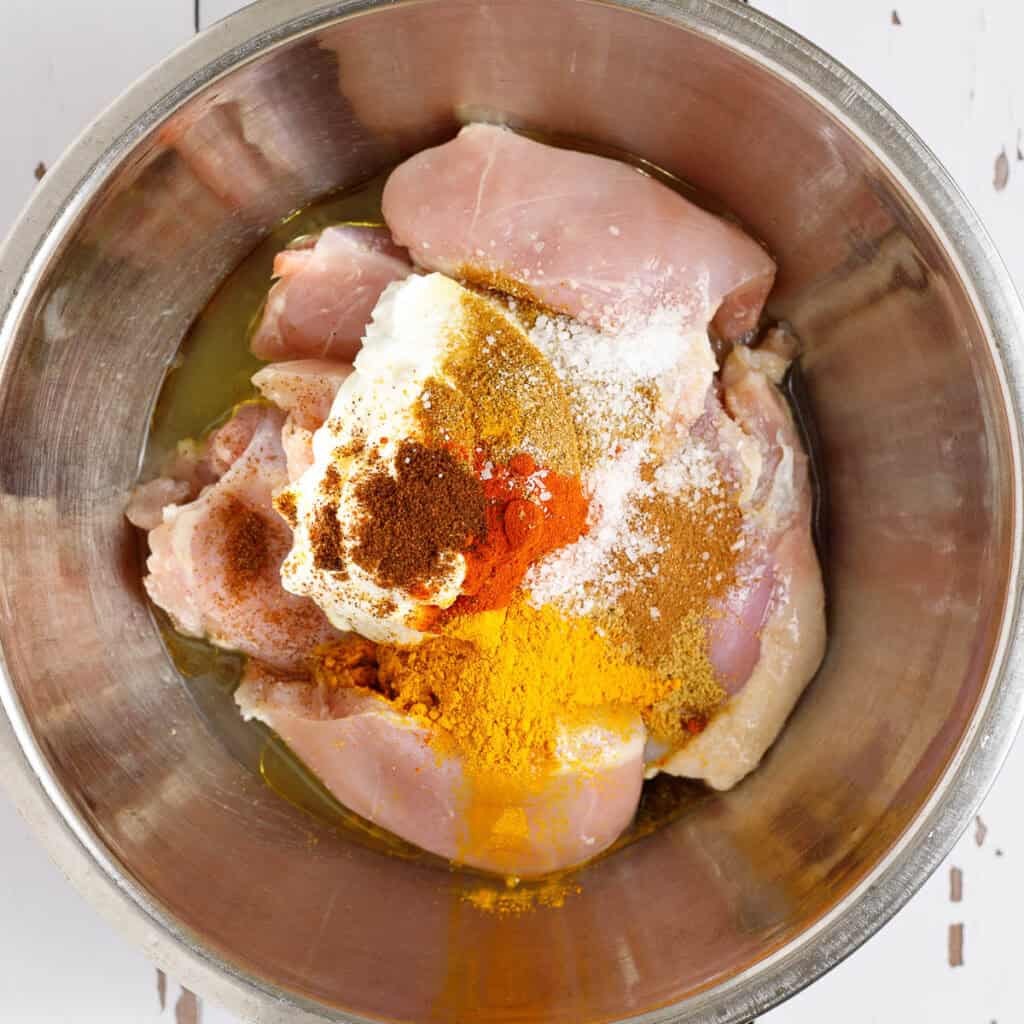 Chicken thighs are shown in a bowl covered with the ingredients for chicken shawarma marinade.