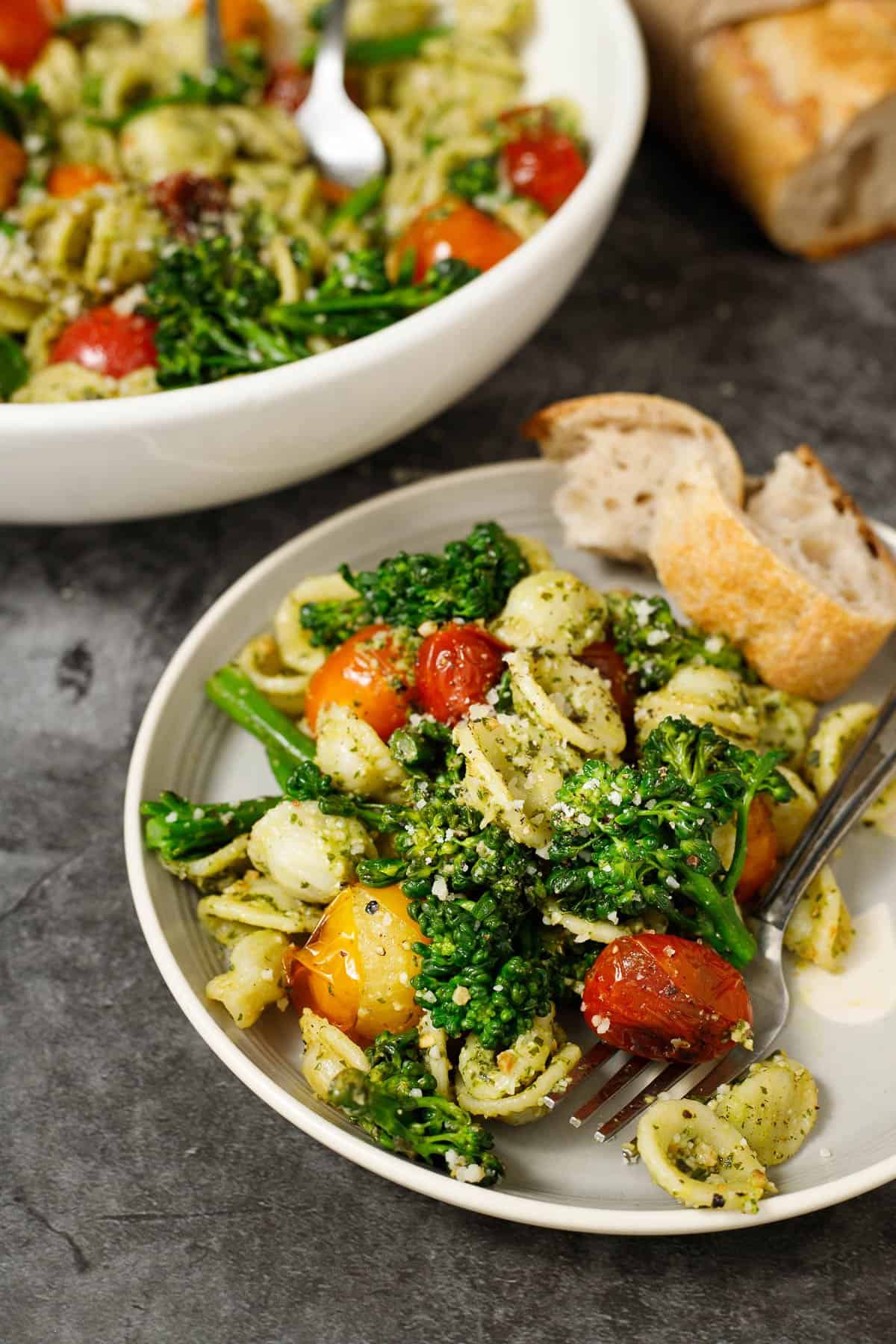 Broccolini pasta with pesto on a plate with a piece of bread. The serving bowl of this dish sits in the background.