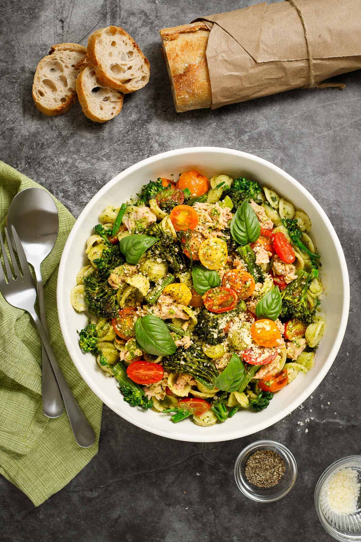 Broccolini pasta recipe in a bowl, accompanied by a napkin and serving utensils. A loaf of bread with several sliced cut off sits nearby.