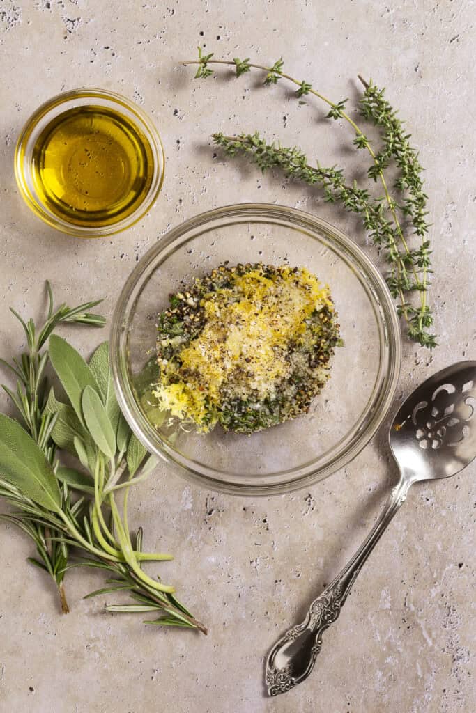An herb rub sits in a bowl surrounded by a smaller bowl of olive oil, a sprig of thyme, and a sprig of rosemary. A spoon sits nearby.
