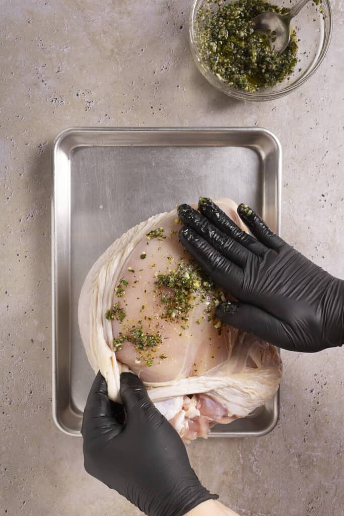 A turkey breast sits on a sheet pan with an herb rub nearby. Someone has gently pulled back the skin of the turkey breast and is putting some of the rub on the meat.