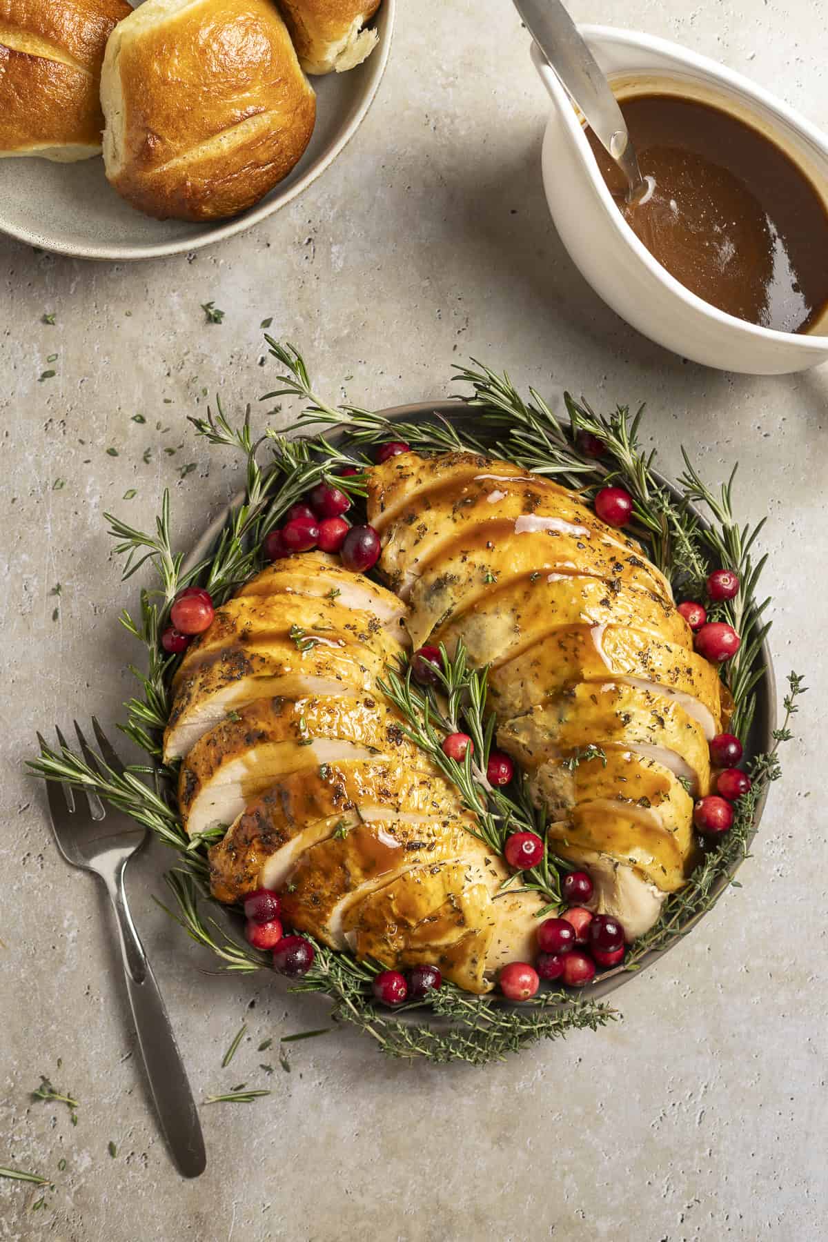 Two turkey breasts with golden brown skin are laid out on a platter and drizzled with balsamic gravy. They are garnished with rosemary and cranberries. A gravy boat sits nearby filled with balsamic gravy, as well as a bowl of rolls.