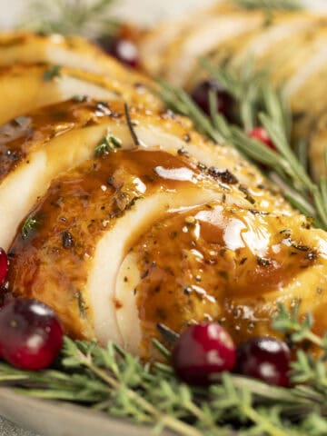 Sliced roasted turkey breast sits on a platter drizzled with balsamic gravy. It is garnished with rosemary and cranberries.