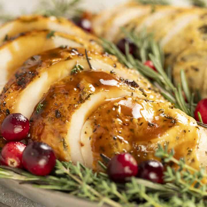 Sliced roasted turkey breast sits on a platter drizzled with balsamic gravy. It is garnished with rosemary and cranberries.