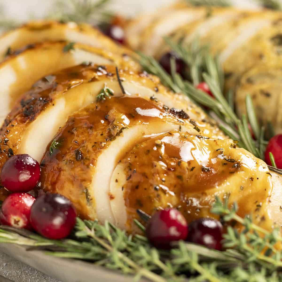 Sliced roasted turkey breast sits on a platter drizzled with balsamic gravy. It is garnished with rosemary and cranberries.