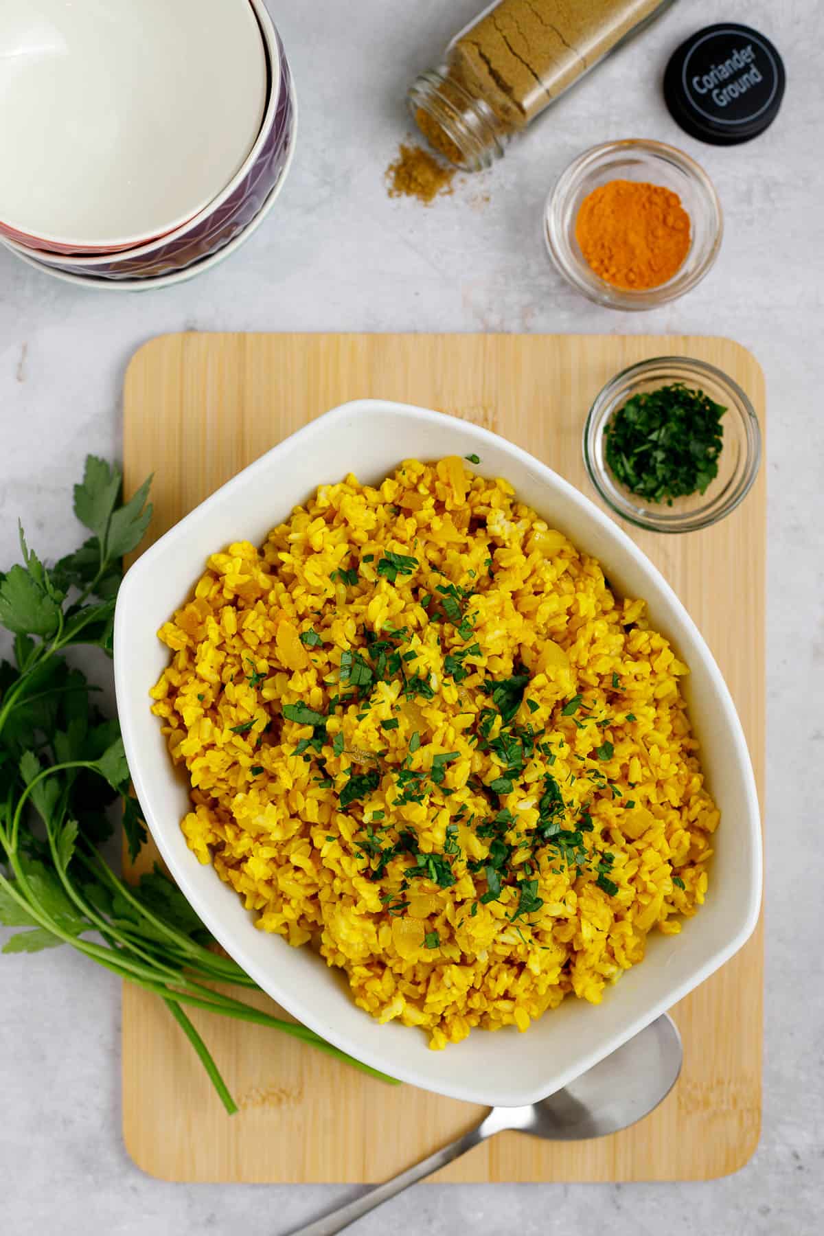 Mediterranean yellow rice in a bowl with a spoon. The bowl sits on a cutting board and is accompanied by spices and herbs. 
