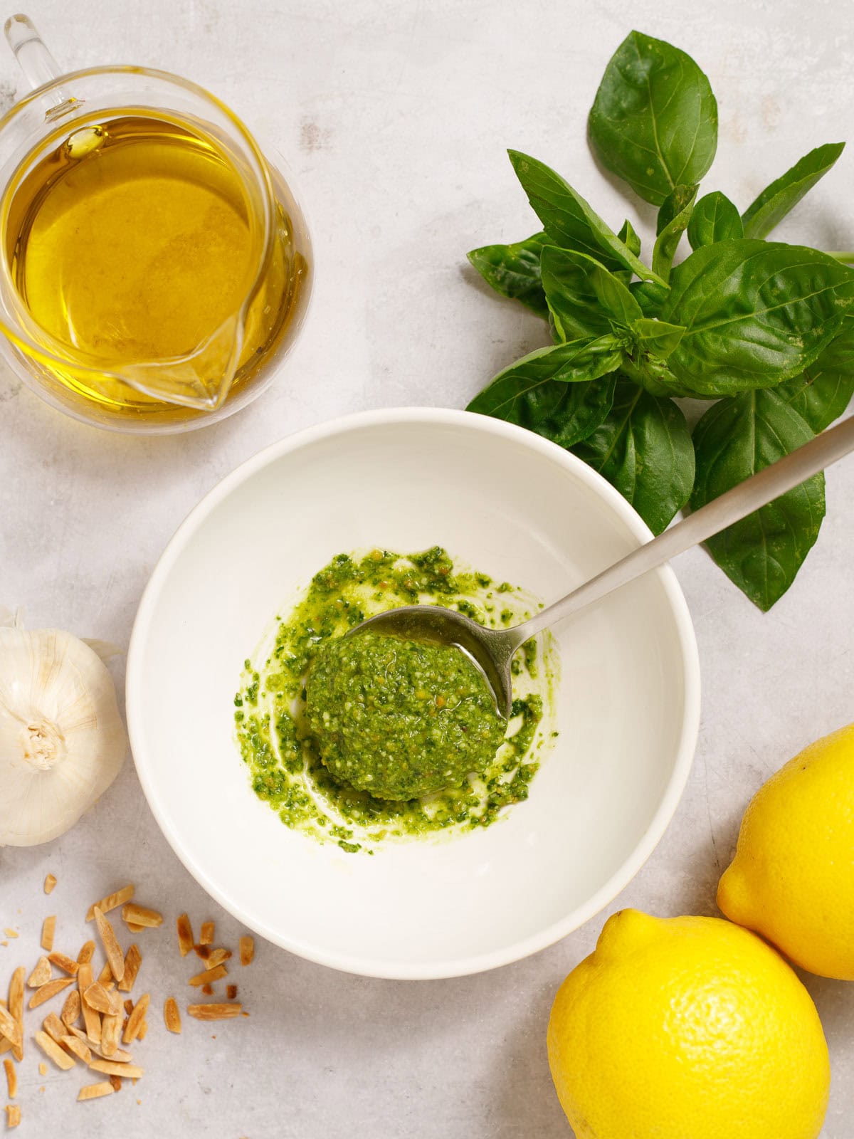 Fresh basil pesto in a white bowl with garlic, lemons, and olive oil on a light background.