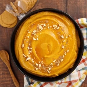 A bowl of pumpkin hazelnut dessert hummus is shown topped with chopped hazelnuts and ground nutmeg. It is served with ginger snap cookies for scooping.