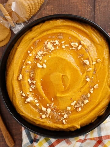 A bowl of pumpkin hazelnut dessert hummus is shown topped with chopped hazelnuts and ground nutmeg. It is served with ginger snap cookies for scooping.