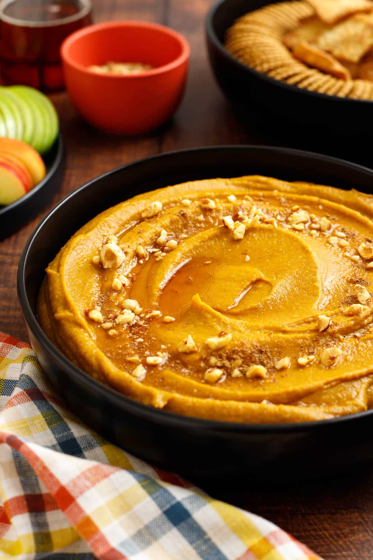 A bowl of pumpkin hazelnut dessert hummus is shown topped with chopped hazelnuts and ground nutmeg. It is served with apple slices, ginger snap cookies, and cinnamon sugar pita chips.
