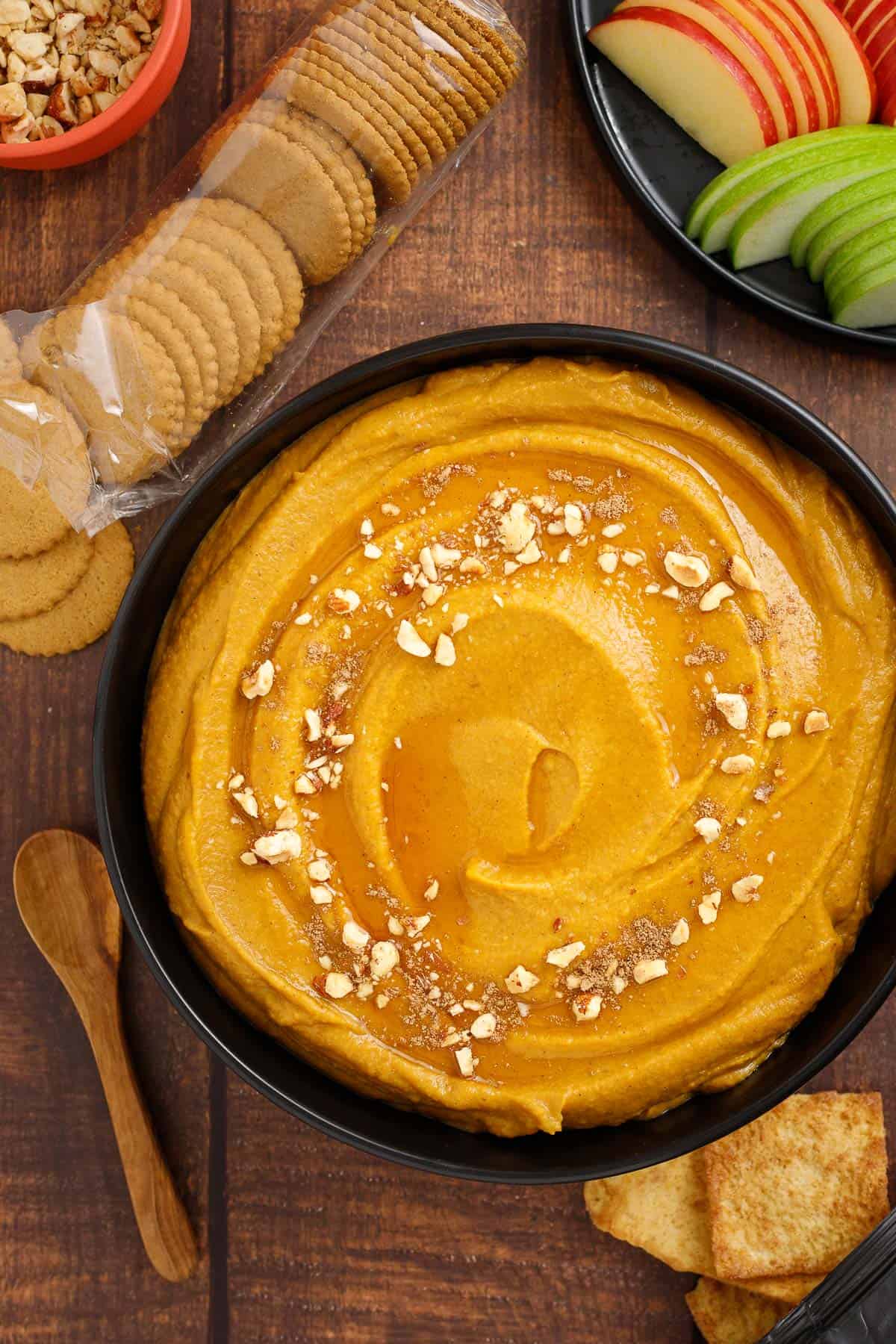 A bowl of pumpkin dessert hummus is surrounded by a spoon, cinnamon pita chips, apple slices, ginger snap cookies, and chopped hazelnuts.