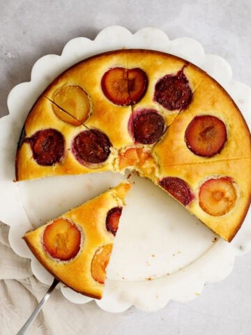 A Mediterranean lemon olive oil cake topped with multi-colored plums sits on a platter, sliced into pieces. Two pieces have already been served, and one sits on a spatula, ready to be served.