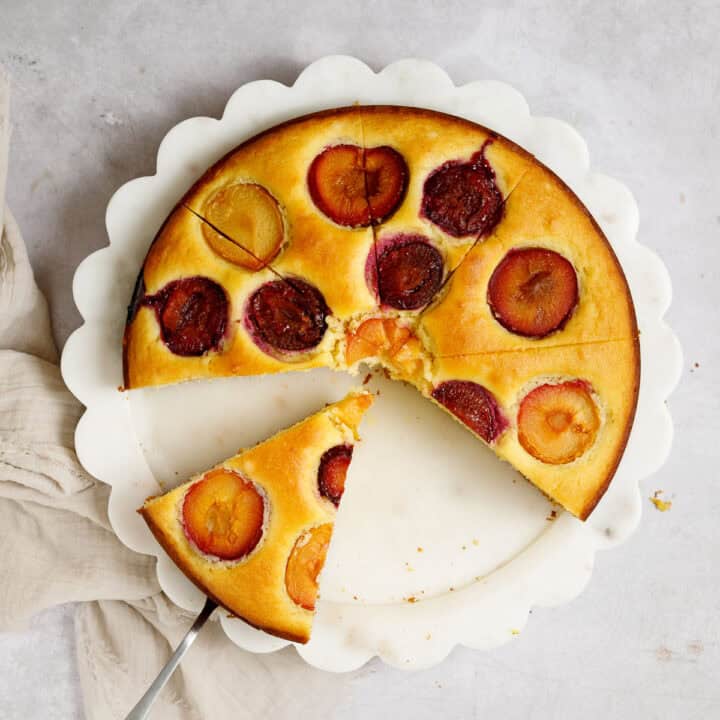 A Mediterranean lemon olive oil cake topped with multi-colored plums sits on a platter, sliced into pieces. Two pieces have already been served, and one sits on a spatula, ready to be served.