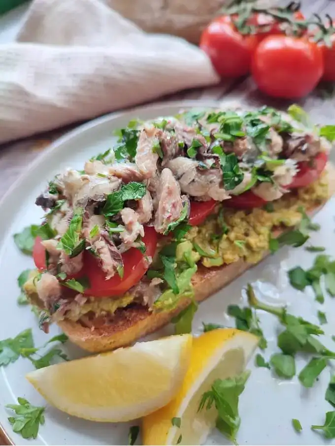 Sardines on toast with avocado and tomato. Topped with herbs and served on a plate with lemon slices.