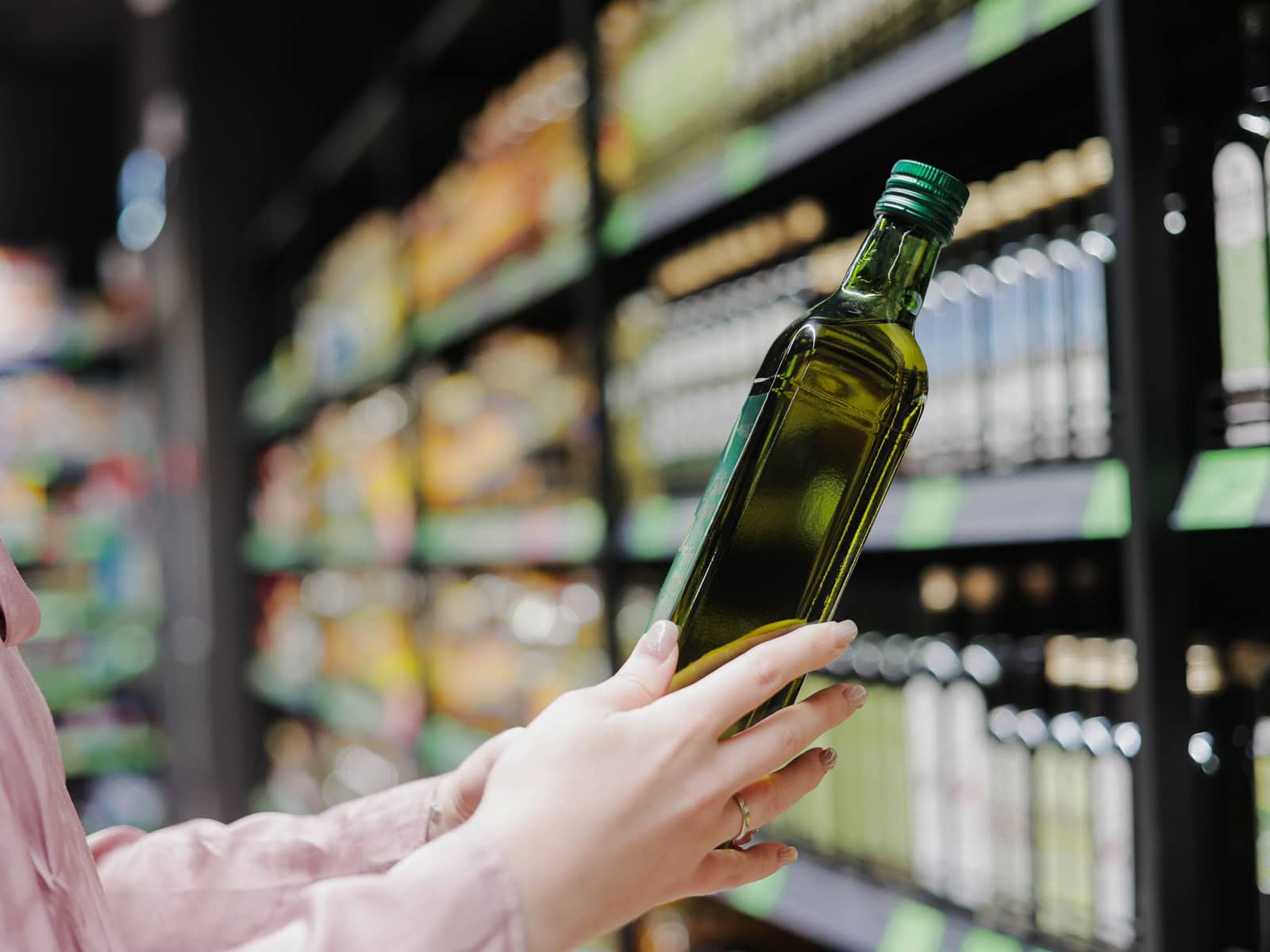 A woman reads the label of a bottle of olive oil in a grocery store, with shelves stocked with olive oil bottles in the background.