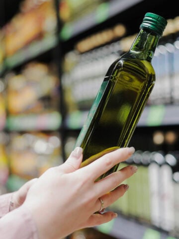 A woman reads a bottle of olive oil in a grocery store with a shelf of olive oil bottles in the background.