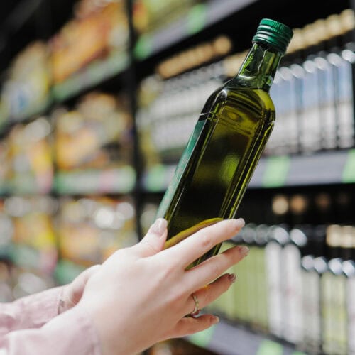 A woman reads a bottle of olive oil in a grocery store with a shelf of olive oil bottles in the background.