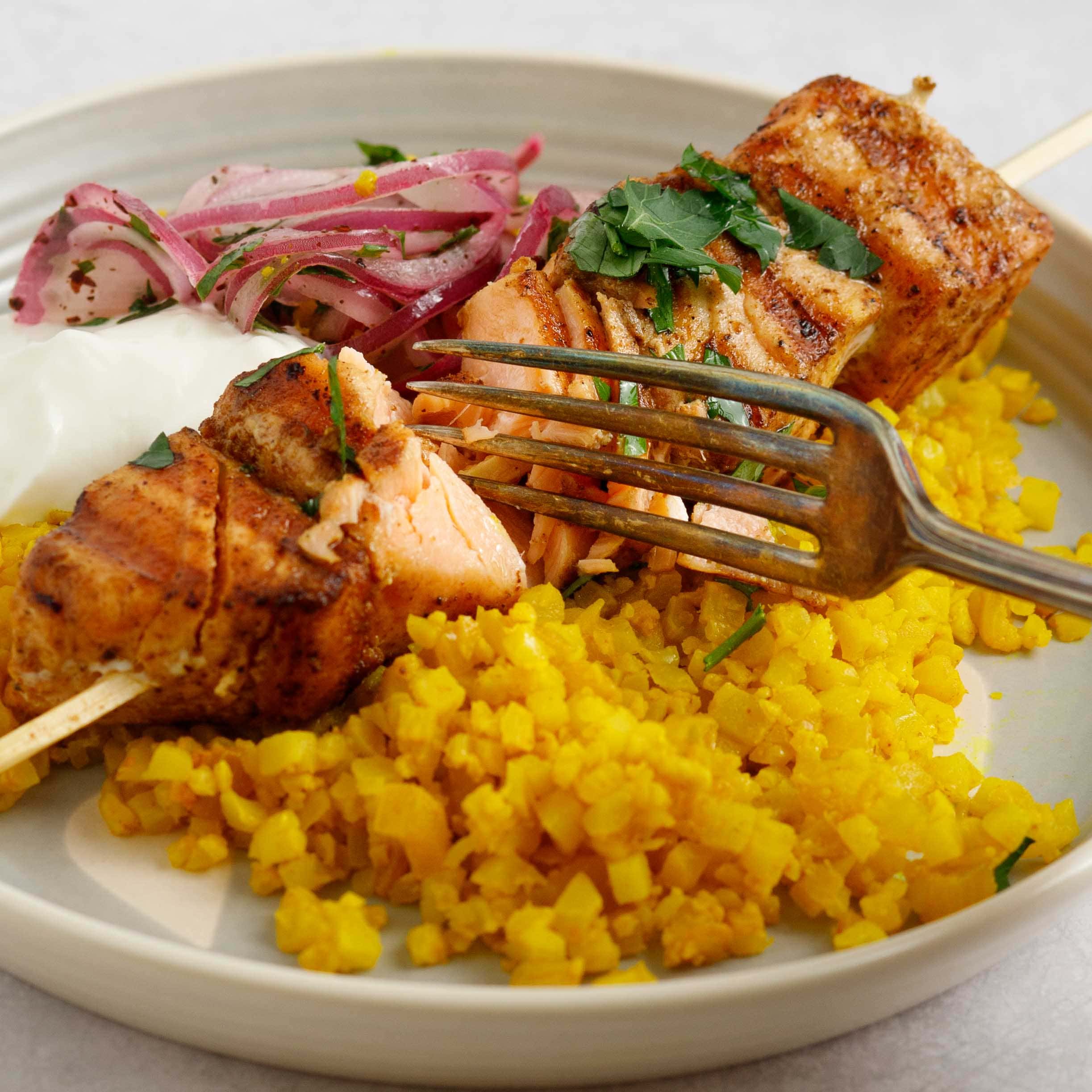 Shawarma salmon skewer on a plate with yellow cauliflower rice being cut with a fork.
