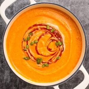 a pot of roasted butternut squash and red pepper soup is garnished with swirls of harissa and tahini and a sprinkling of roasted pepitas.