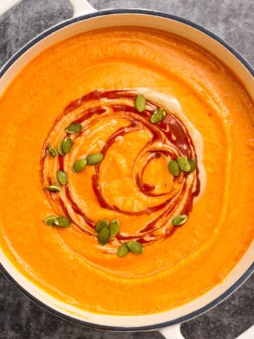 a pot of roasted butternut squash and red pepper soup is garnished with swirls of harissa and tahini and a sprinkling of roasted pepitas.