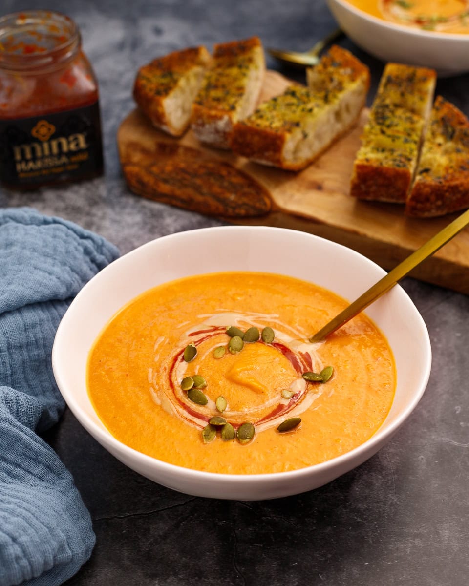 butternut squash and red pepper soup in a bowl garnished with tahini and harissa. A jar of harissa sits in the back with a cutting board with toasted bread slices.
