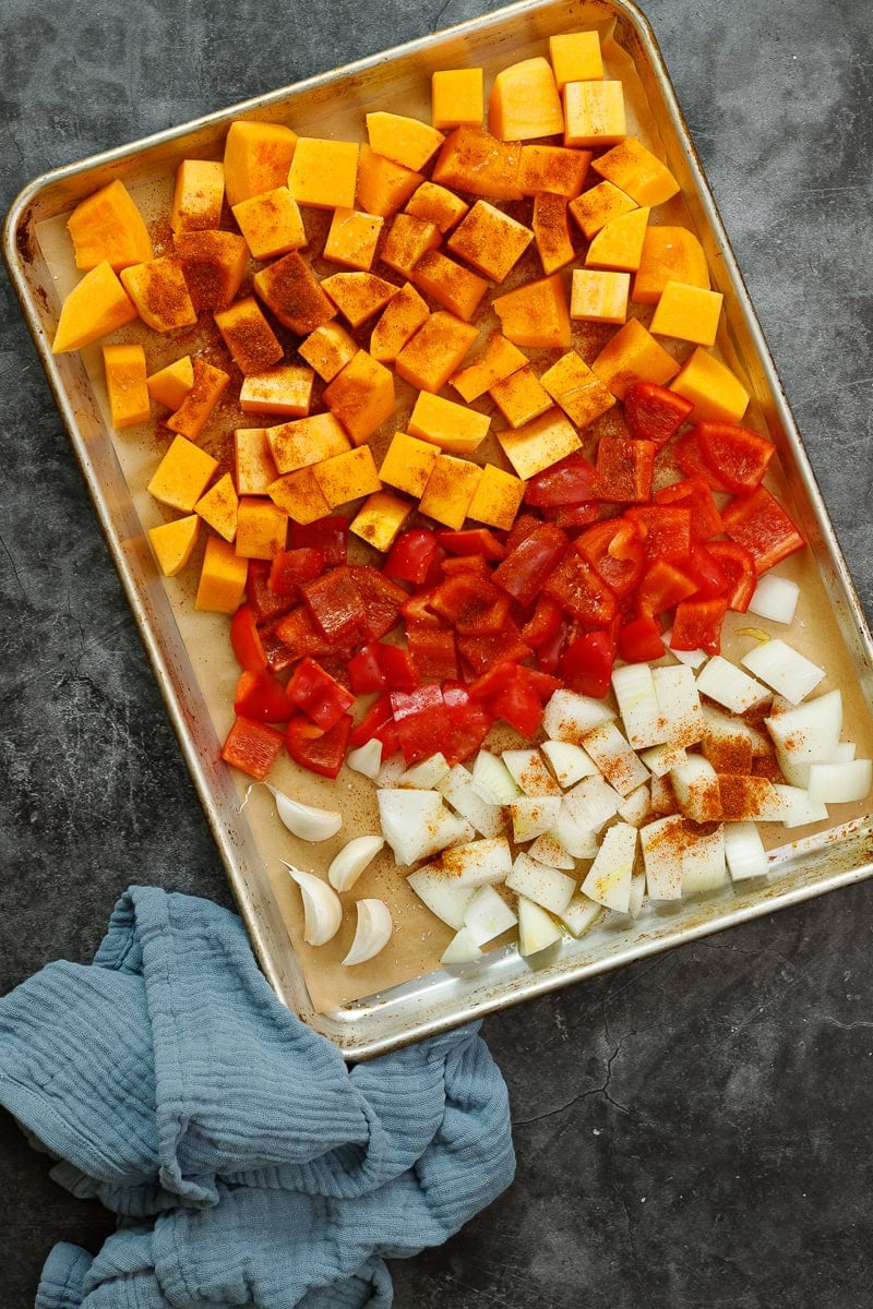 Chopped butternut squash, red bell pepper, and onion on a sheet pan drizzled with olive oil, salt, and harissa seasoning.