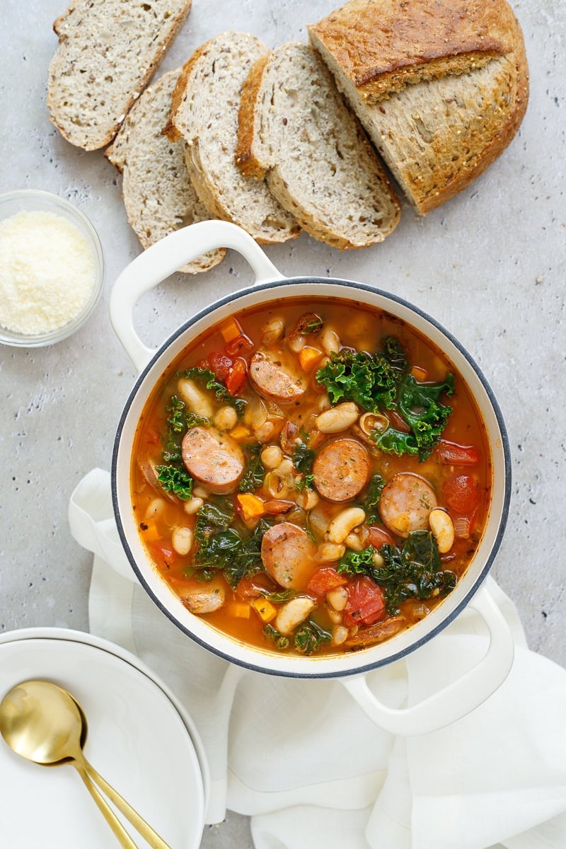 Italian sausage and kale soup with white beans in a pot on a stone surface. It is surrounded by sliced bread, two bowls and spoons, grated parmesan cheese, and a white napkin.